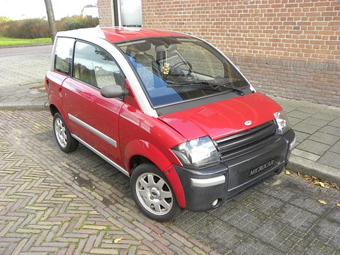 Red small hatchback with a slatted black nose parked on paving by a brick wall. Opens a larger view