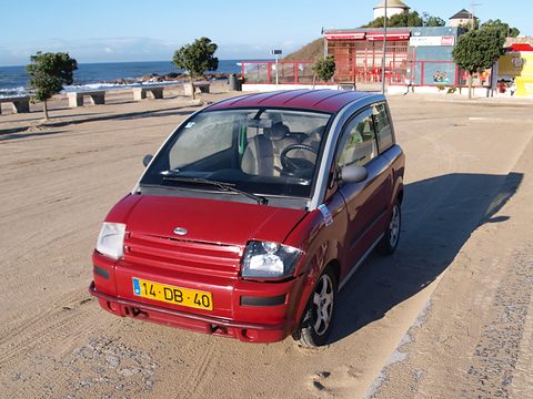 Red microcar with a Portuguese yellow plate parked on sand, the sea and a promenade behind. Opens a larger view