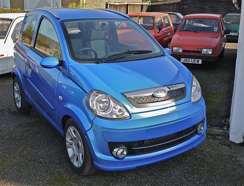 Bright blue small hatchback with swept headlamps and chrome grille bar, parked by other cars.