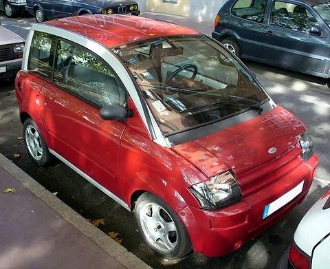 Red microcar with a silver roof frame and slatted nose parked beside a kerb. Opens a larger view