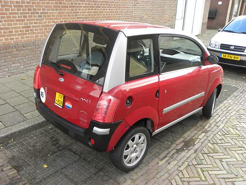 Red microcar with silver roof pillars and a 45 disc, seen from the rear on brick paving. Opens a larger view