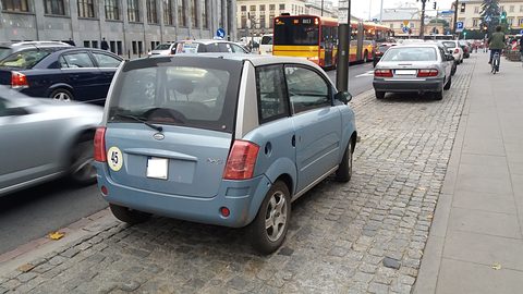 Pale blue microcar seen from the rear on cobbles beside a busy street with a tram stop. Opens a larger view