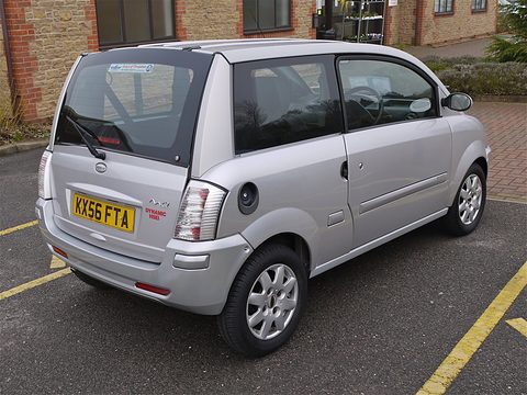 Rear of a silver microcar with tall tail lamps parked in front of a brick building. Opens a larger view