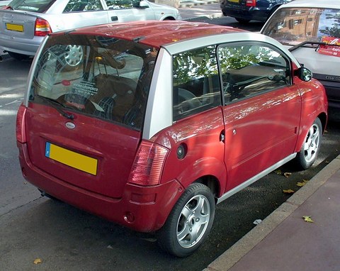 Rear of a red microcar with silver roof pillars parked at a kerb in dappled sun. Opens a larger view