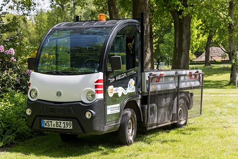 Black and white small electric utility truck with an amber beacon and cage body on a lawn under trees. Opens a larger view