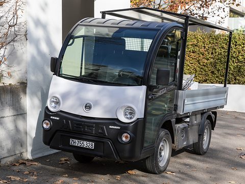 Black and white small electric utility truck with round headlamps and an open-frame bed beside a white wall. Opens a larger view