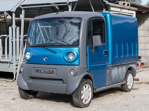 Blue and grey small electric box van with round headlamps and a MEGA grille parked in a yard. Opens a larger view