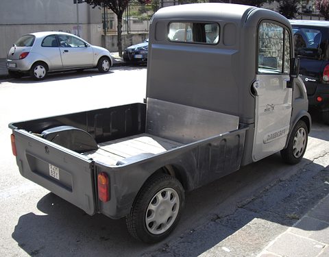 Rear of a grey small pickup with a black load bed and a tall cab, parked beside a dark car. Opens a larger view