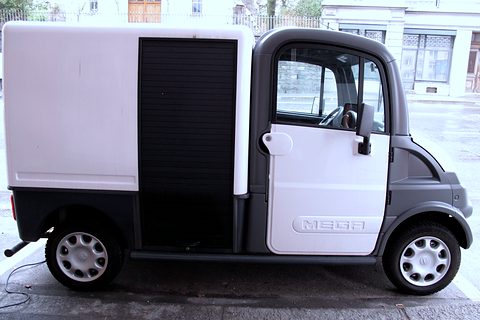 Side view of a white and grey small electric van with a box body and roller shutter on a wet road. Opens a larger view