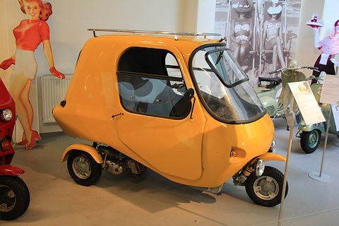A rounded orange enclosed three-wheeler with a curved windscreen and a chrome roof rack, a scooter behind it. Opens a larger view