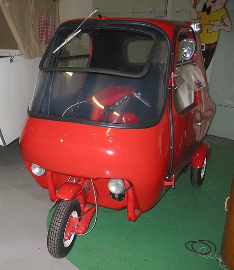 Small red closed cabin three-wheeler with a big curved windscreen, on a green floor.
