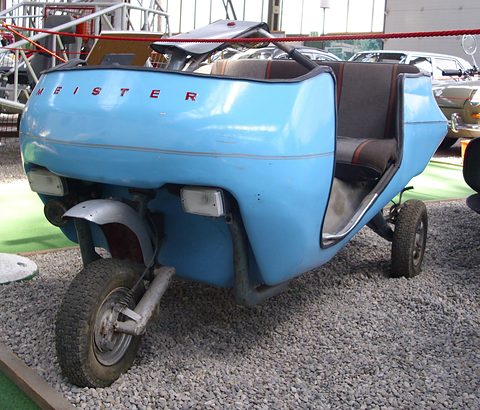 Pale blue open microcar with a dark seat, MEISTER lettering on the tail, on a gravel museum bed. Opens a larger view