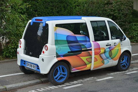 White tall microvan with swirling multicoloured graphics and blue wheels parked at a kerb by a hedge. Opens a larger view
