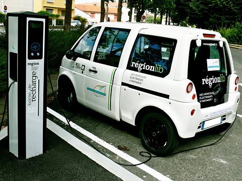 White tall microvan lettered régionlib parked beside a tall black and white charging pillar. Opens a larger view