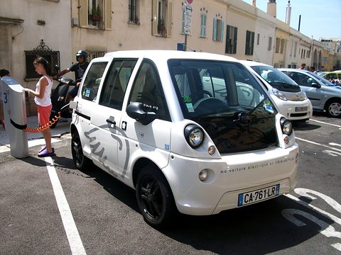 White tall microvan connected by an orange cable to a charging post, people and scooters on the street behind. Opens a larger view