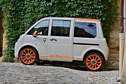 White tall microvan with orange wheels and miAmore script, parked on cobbles against an ivy-covered wall. Opens a larger view