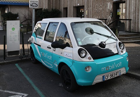 Turquoise and white tall microvan lettered mobilivolt parked at a charging bay by a building. Opens a larger view