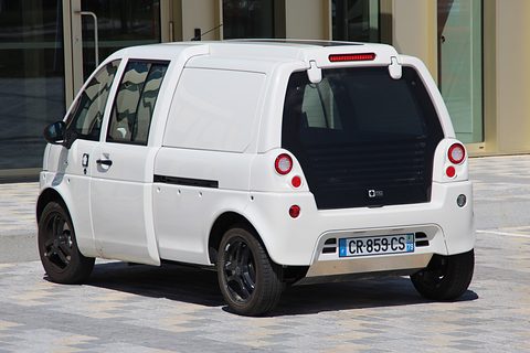 Rear of a white tall microvan with windowless rear sides, round tail lamps and black wheels on paving. Opens a larger view