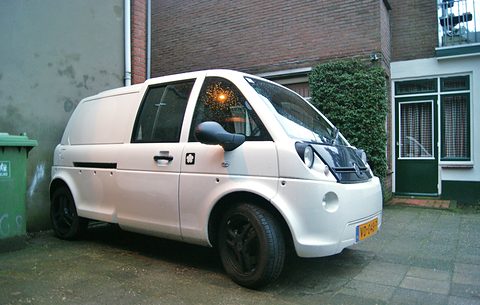 Side view of a white tall microvan with a sliding door and black wheels on brick paving by a house. Opens a larger view