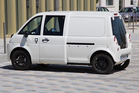 Side view of a white tall microvan with a blanked rear panel and black wheels on stone paving. Opens a larger view