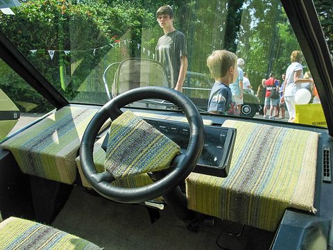 Woven striped seats and a black steering wheel seen through an open side, a garden behind. Opens a larger view