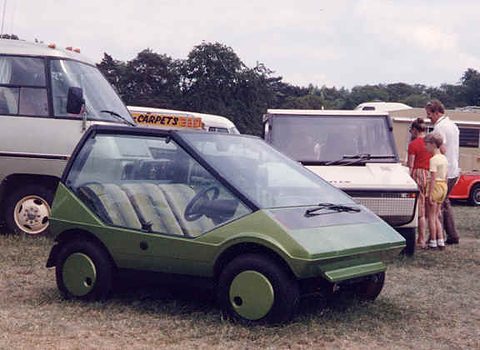 Olive-green wedge-shaped concept car on grass, a family looking at a white car behind. Opens a larger view