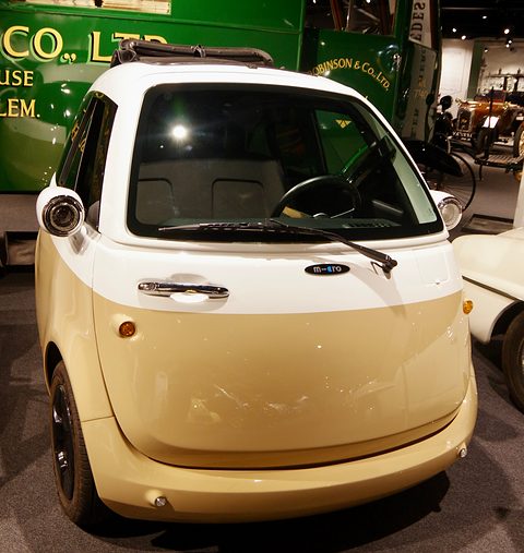Cream and white bubble-shaped car seen head-on, its whole front forming the door, beside a green vintage lorry in a museum hall. Opens a larger view