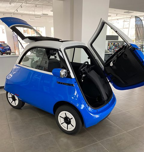 Blue and white bubble car with its whole front swung open and its rear hatch raised, on a tiled showroom floor.