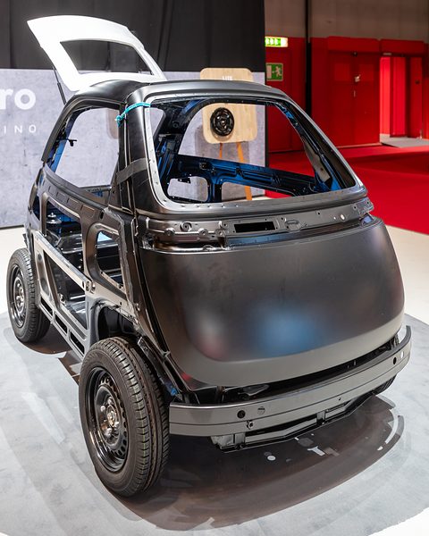 Black unpainted car body with welded pressings and a front door frame on wheels, on a show floor. Opens a larger view