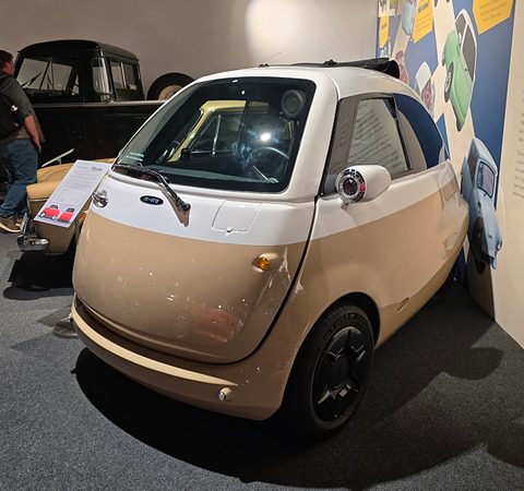 Cream and white bubble car on a carpeted museum floor beside display panels, another small car behind. Opens a larger view
