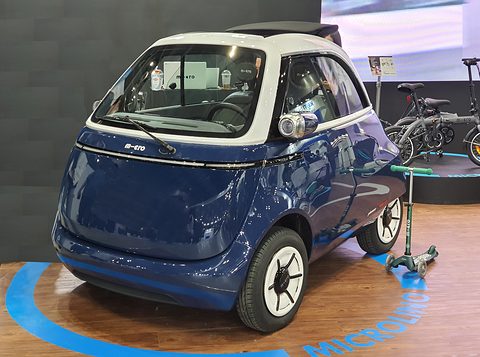 Blue and white bubble car on a wooden stand with a Microlino ring, folding bicycles behind. Opens a larger view