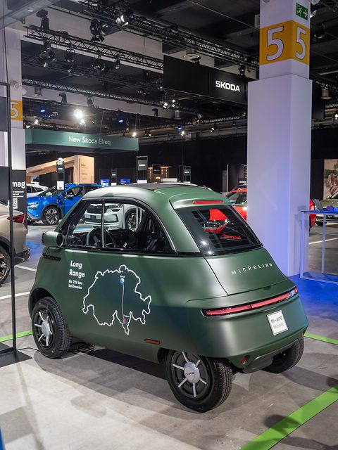 Rear of a matt-green bubble car with a white outline map of Switzerland on its side, on a show floor. Opens a larger view
