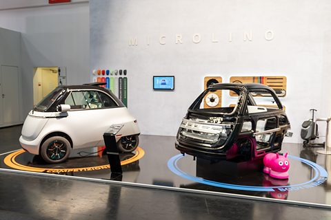 White bubble car and a black bare body structure on round turntables in front of a white Microlino wall.