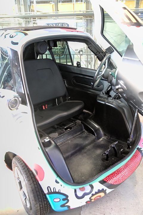 Black bench seat and bare floor inside a small car, seen through a wide front opening edged with patterned panels. Opens a larger view