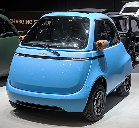 Light-blue bubble car with orange lamp pods and a dark roof on a show floor, a bare body shell behind.