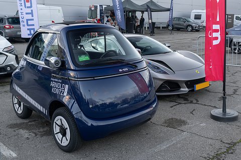 Navy bubble car with dealer lettering parked on tarmac beside a silver sports car, gazebos and flags behind. Opens a larger view