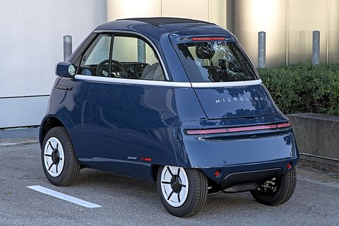 Rear of a navy bubble car with white wheels and a tail-light strip, parked on tarmac by a building. Opens a larger view
