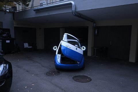 Blue and white bubble car with its whole front swung open as a door, in a dim concrete courtyard by garages. Opens a larger view