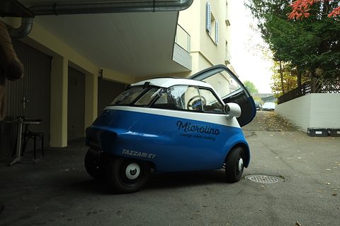 Side view of a blue and white bubble car with Microlino and Tazzari EV lettering, its front door raised.