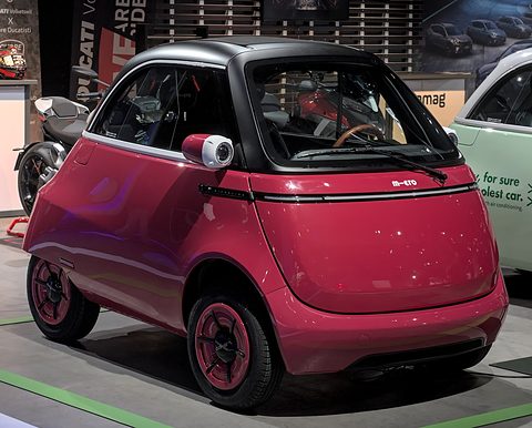 Crimson bubble car with a black roof and crimson wheels on a show floor, a motorcycle behind. Opens a larger view