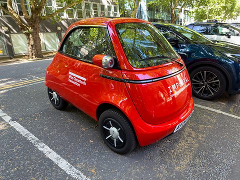 Red bubble car with lettering on its side parked in a street bay beside a dark SUV, plane trees behind. Opens a larger view