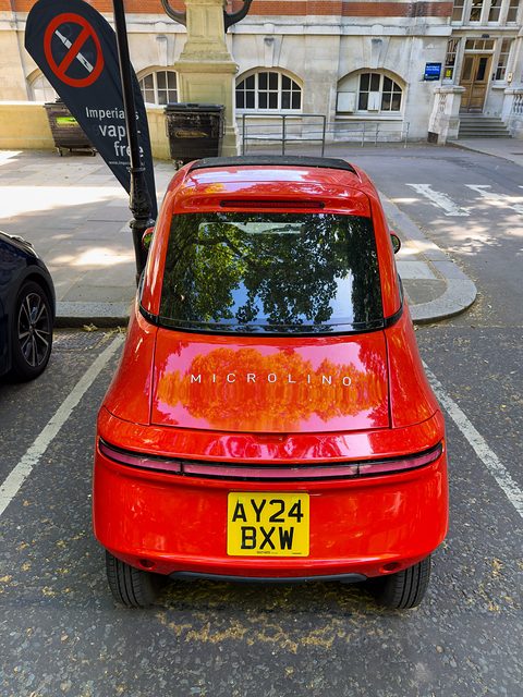 Rear of a red bubble car with a full-width tail-light strip and a British plate, parked on a street. Opens a larger view
