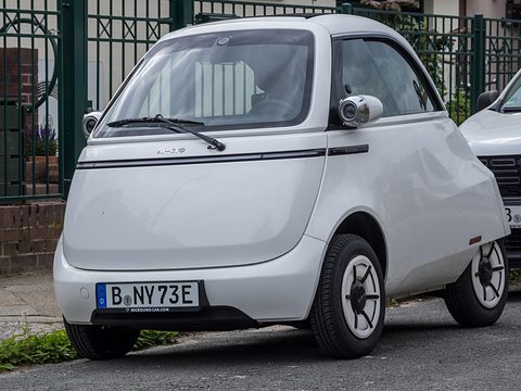 White bubble car with a Berlin plate parked by a green iron fence and a brick wall. Opens a larger view
