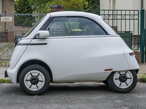 Side view of a white bubble car with white wheels parked on a pavement by a wire fence. Opens a larger view
