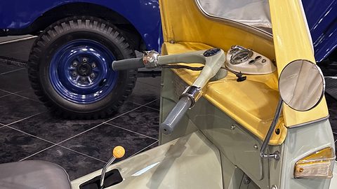Grey motorcycle-style handlebars with a twist grip and a small round speedometer inside a cream and pale green cab.
