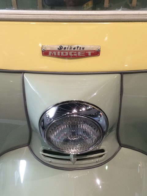 Chrome-rimmed round headlamp in a pale green nose below a cream panel carrying a red Daihatsu Midget badge. Opens a larger view
