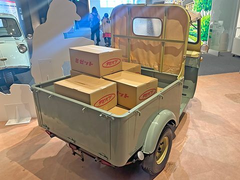 Three-wheeled pickup seen from behind with cardboard boxes in its drop-side bed and a canvas cab top. Opens a larger view