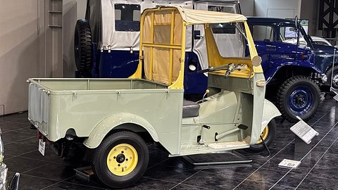 Pale green three-wheeled pickup from behind with a drop-side load bed, yellow wheels and a canvas cab top. Opens a larger view