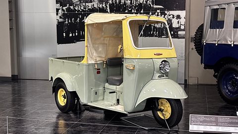 Pale green and cream three-wheeler with a canvas top and single front wheel in front of a large black-and-white crowd mural. Opens a larger view