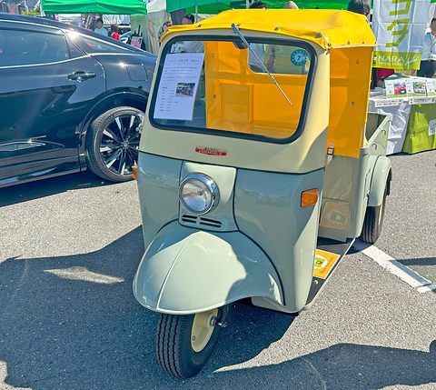 Pale green and cream three-wheeled pickup with a yellow canvas cab and one headlamp parked on tarmac beside a black car. Opens a larger view